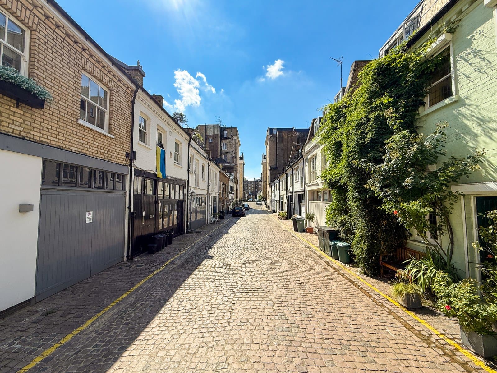 Cobblestone street in a charming London neighbourhood with colourful buildings and lush greenery under a bright blue sky.