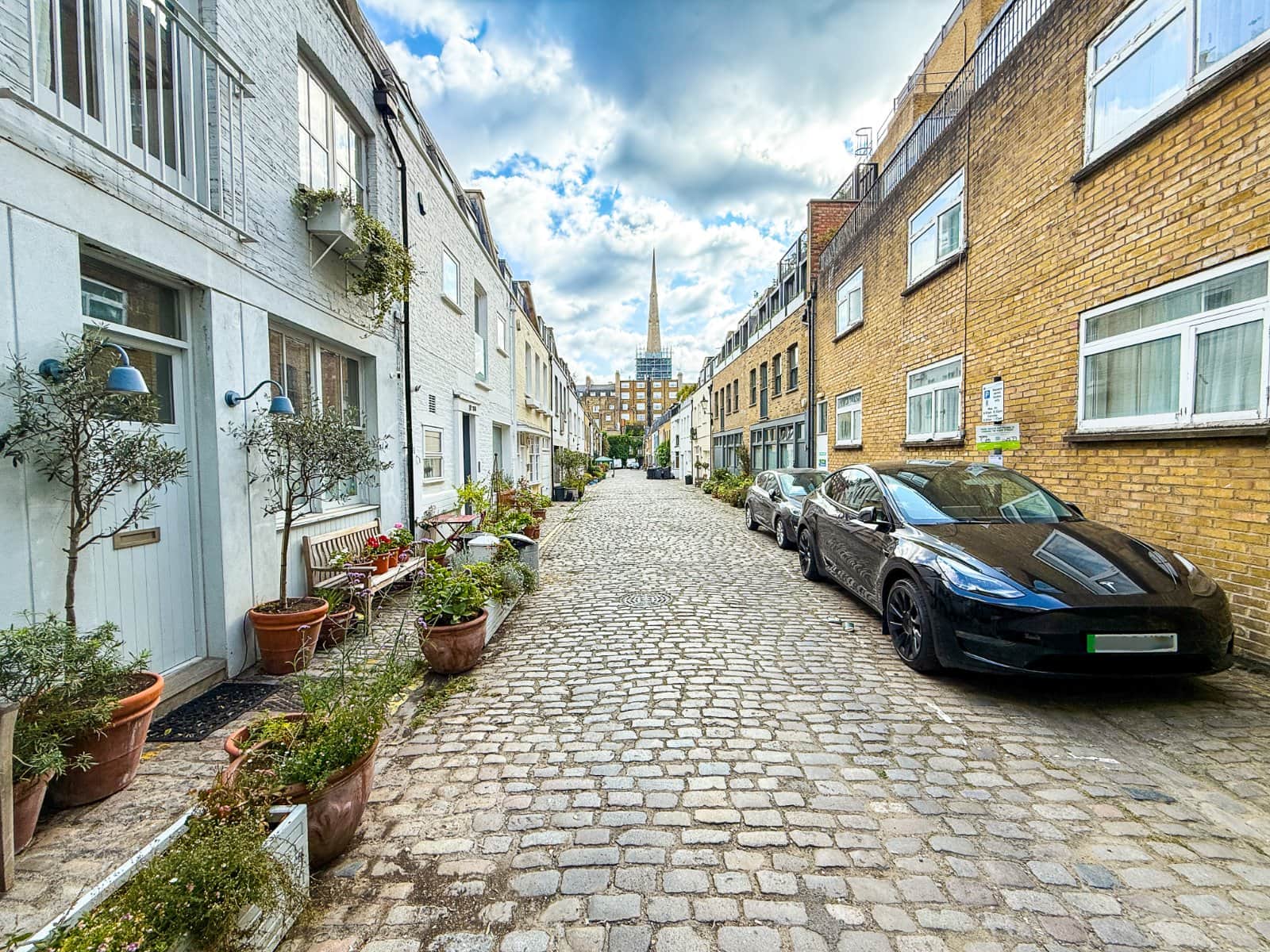 Charming cobbled residential street in London with modern homes, colourful plants, and luxury cars, featuring a prominent church spire in the background.