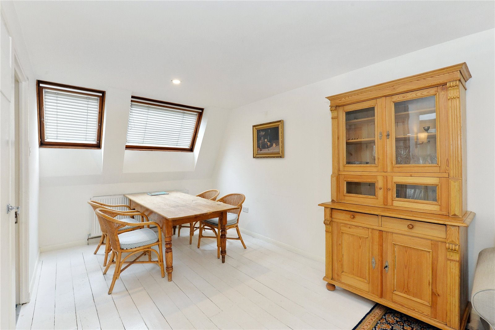 Rustic wooden dining table with rattan chairs in bright attic space, two skylight windows, white walls, classic wooden cabinet.