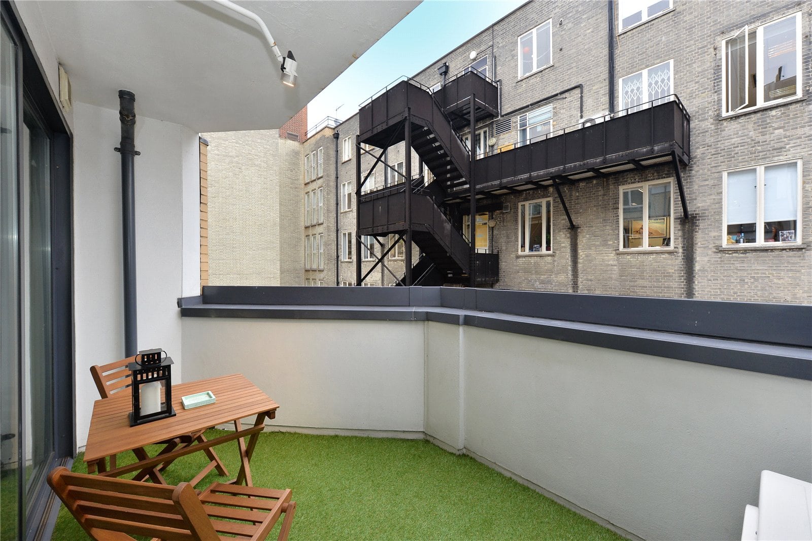 Cozy balcony with artificial grass, wooden furniture, and black lanterns overlooking urban brick buildings.