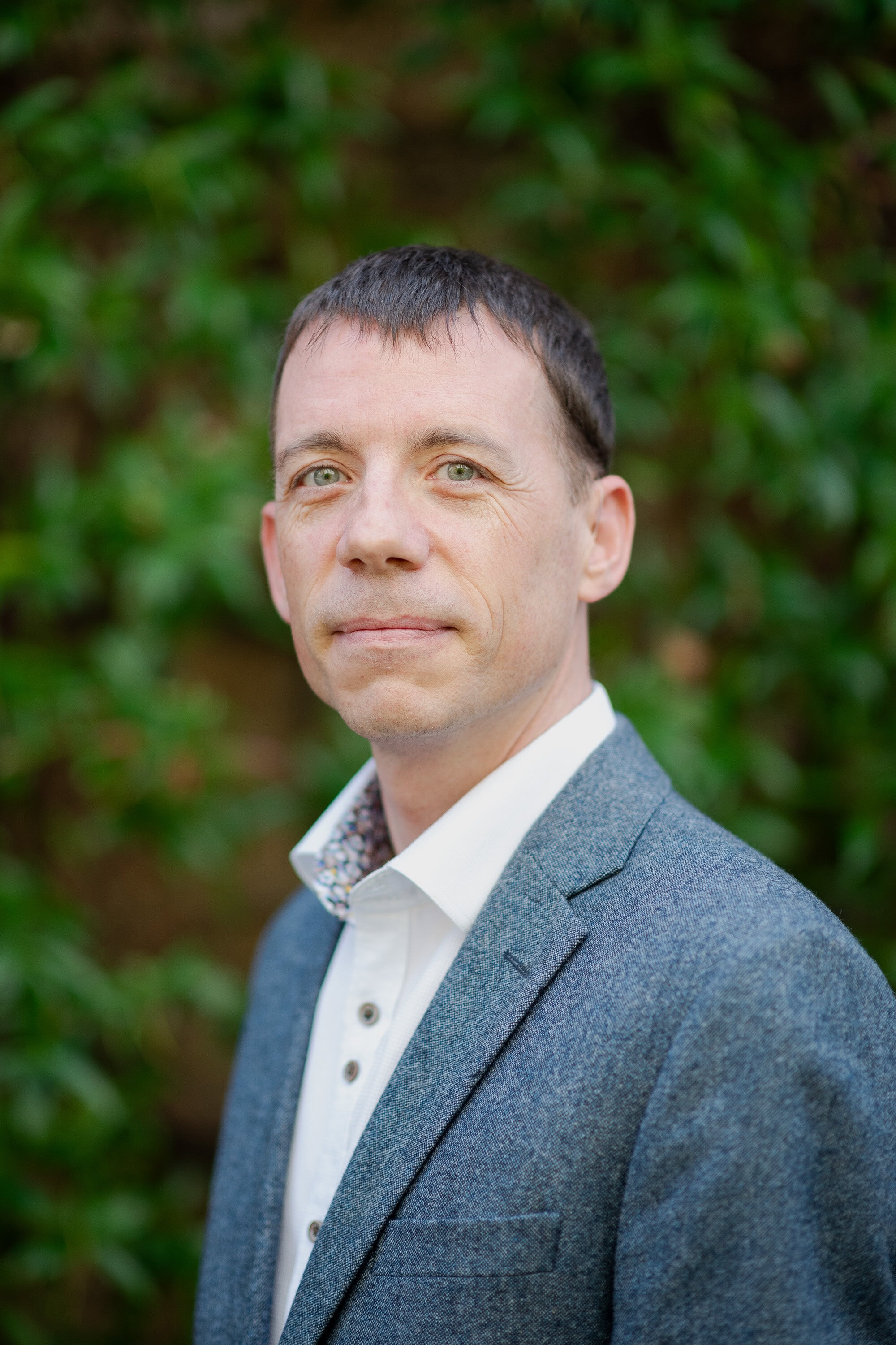 Professional estate agent portrait outside, London property expert, Lurot Brand real estate agent, green shrub background, business attire.