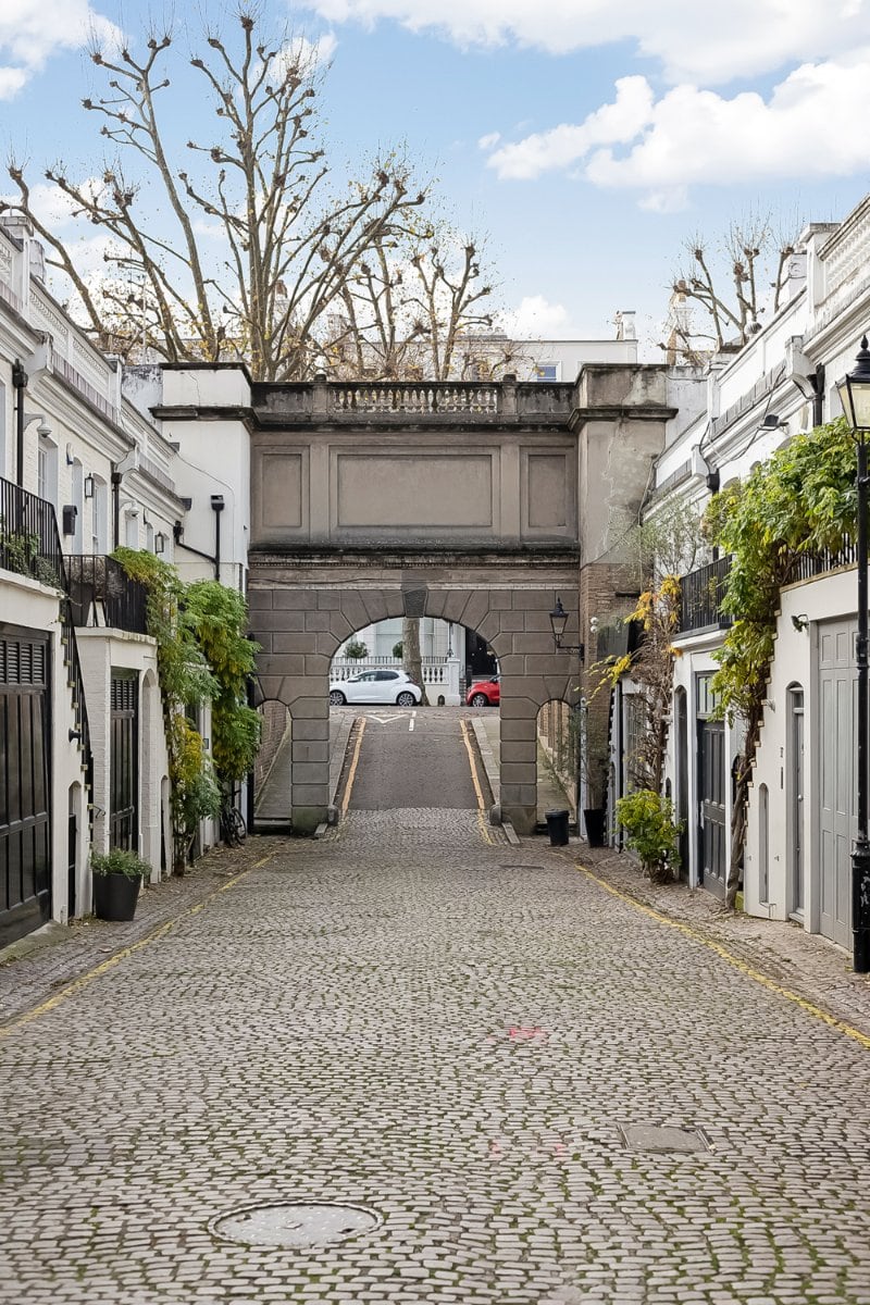 Charming cobbled street leading to a historic archway in a London neighbourhood. Elegant white terraced houses with greenery and vintage street lamps.
