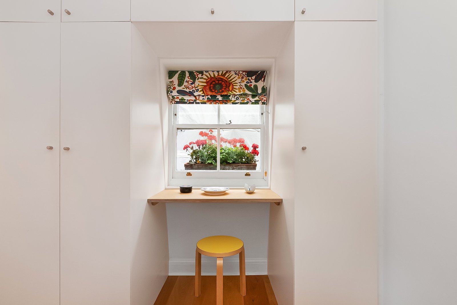 Bright white built-in wardrobes surrounding a small window breakfast nook with colourful floral blinds and a wooden stool.