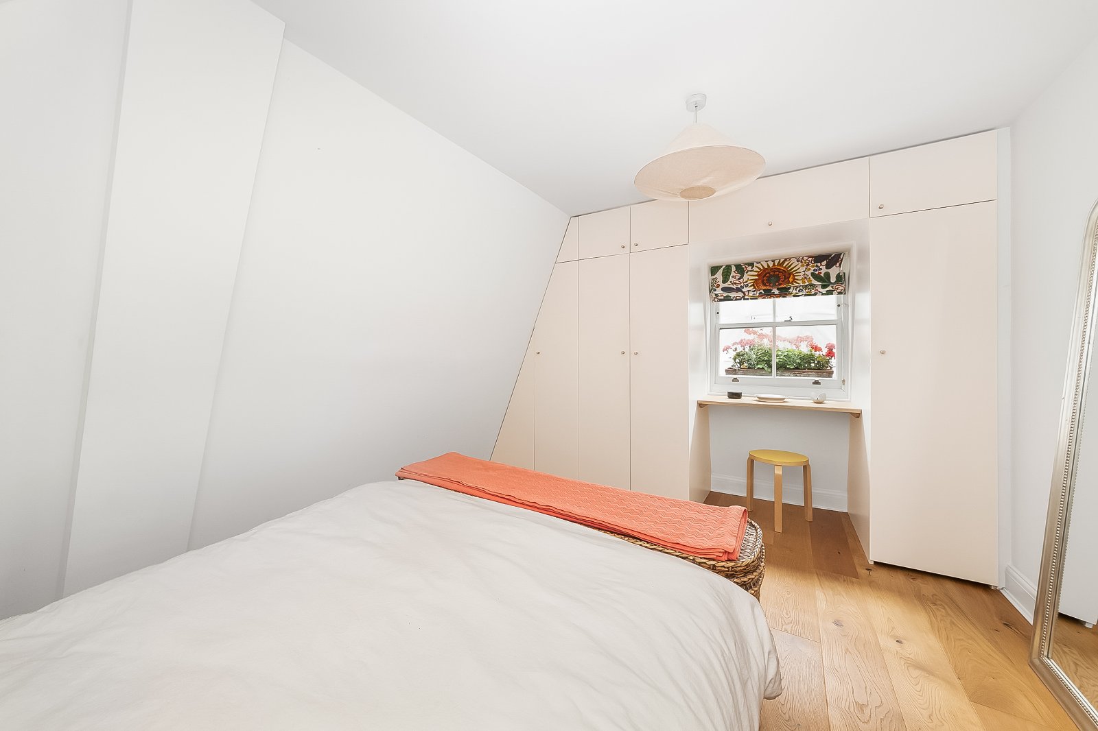 Bright bedroom with built-in white wardrobe, wooden flooring, and a small desk under a window with colourful floral curtain.