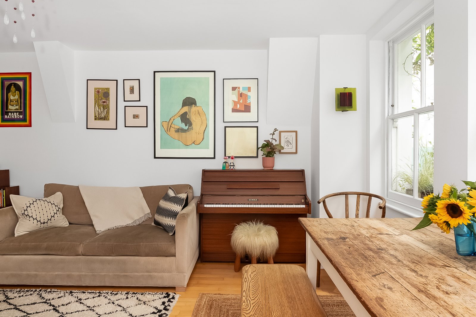 Vibrant living room interior with artful gallery wall, beige sofa, vintage piano, and large window with natural sunlight.