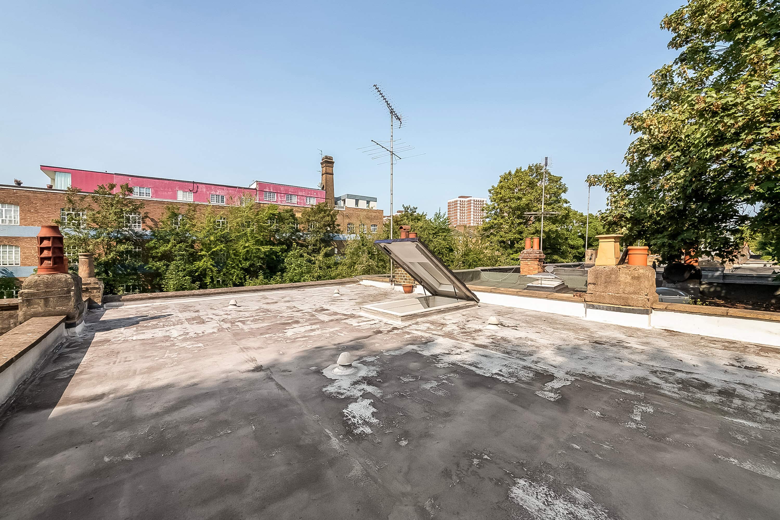 Urban rooftop with skylight and chimney vents in a cityscape, featuring trees and multistory buildings under a clear sky.