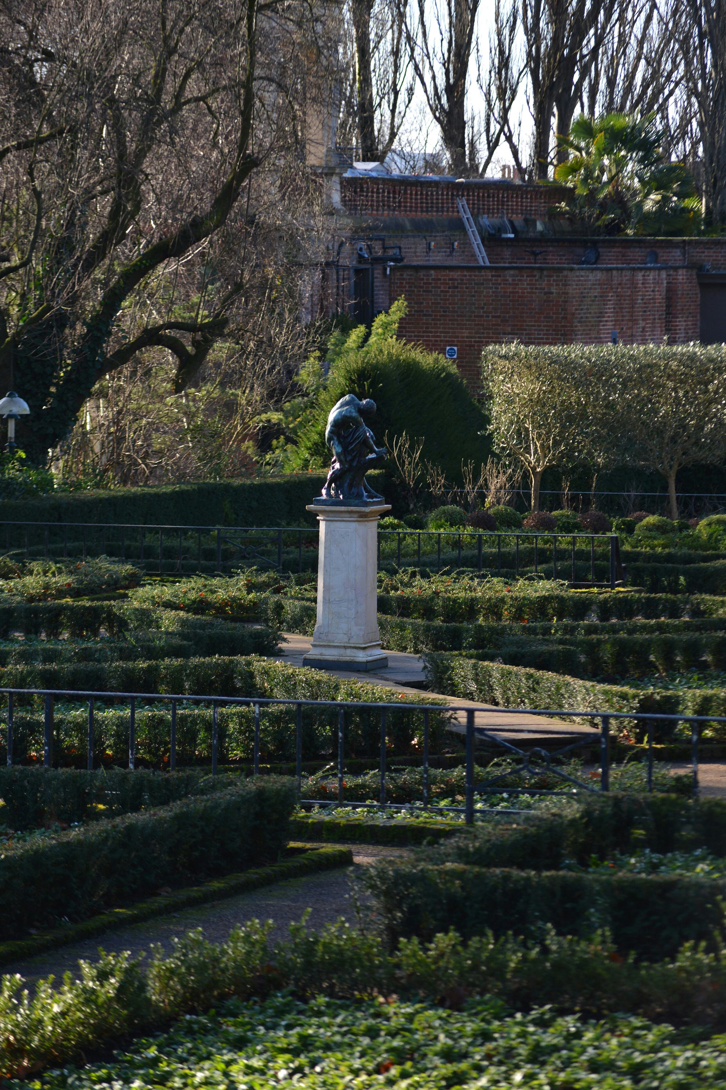 Bust statue in a formal garden with topiary and hedges, historic brick wall in the background.