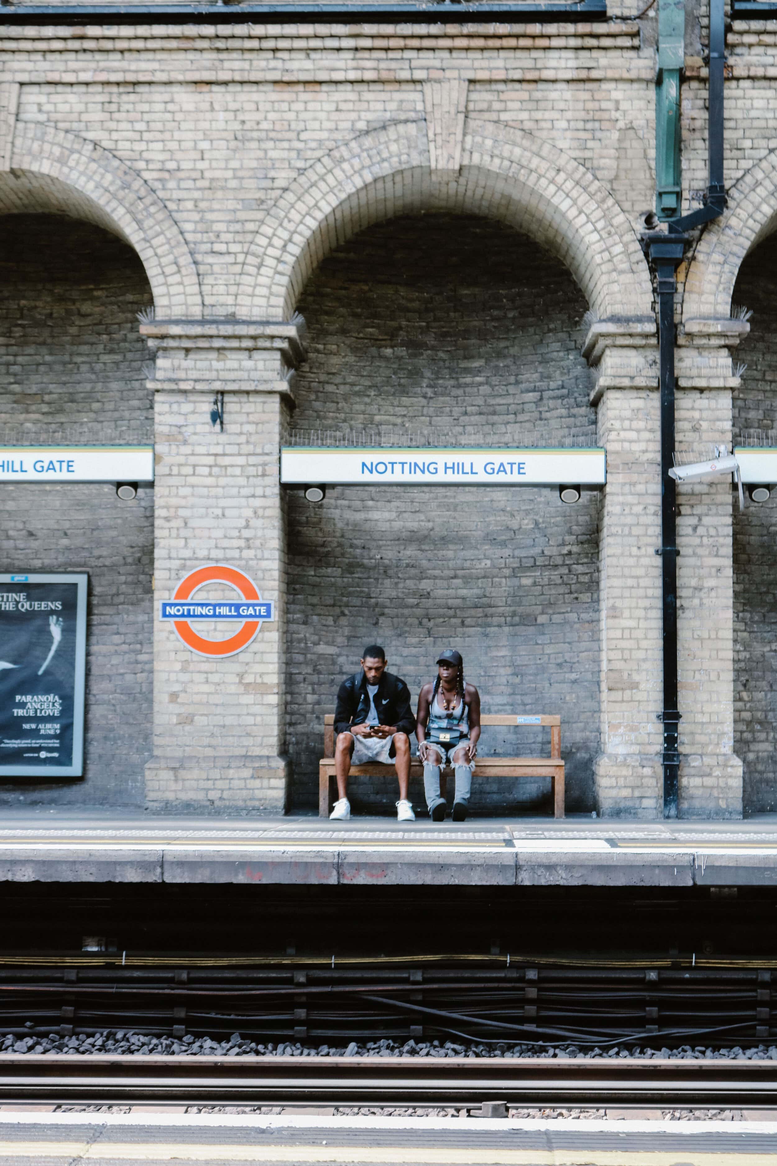 Two people sitting on a bench at Notting Hill Gate station platform.